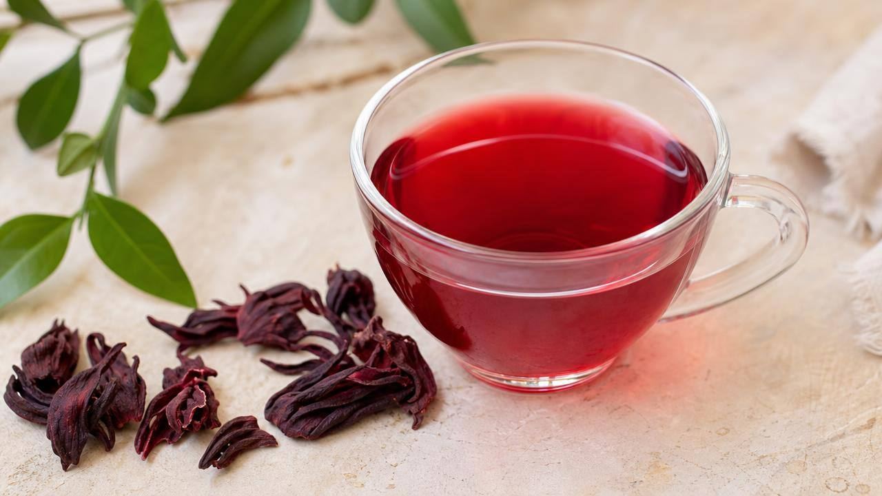 Xícara de chá de hibisco vermelho ao lado de cálices secos de hibisco sobre uma mesa clara