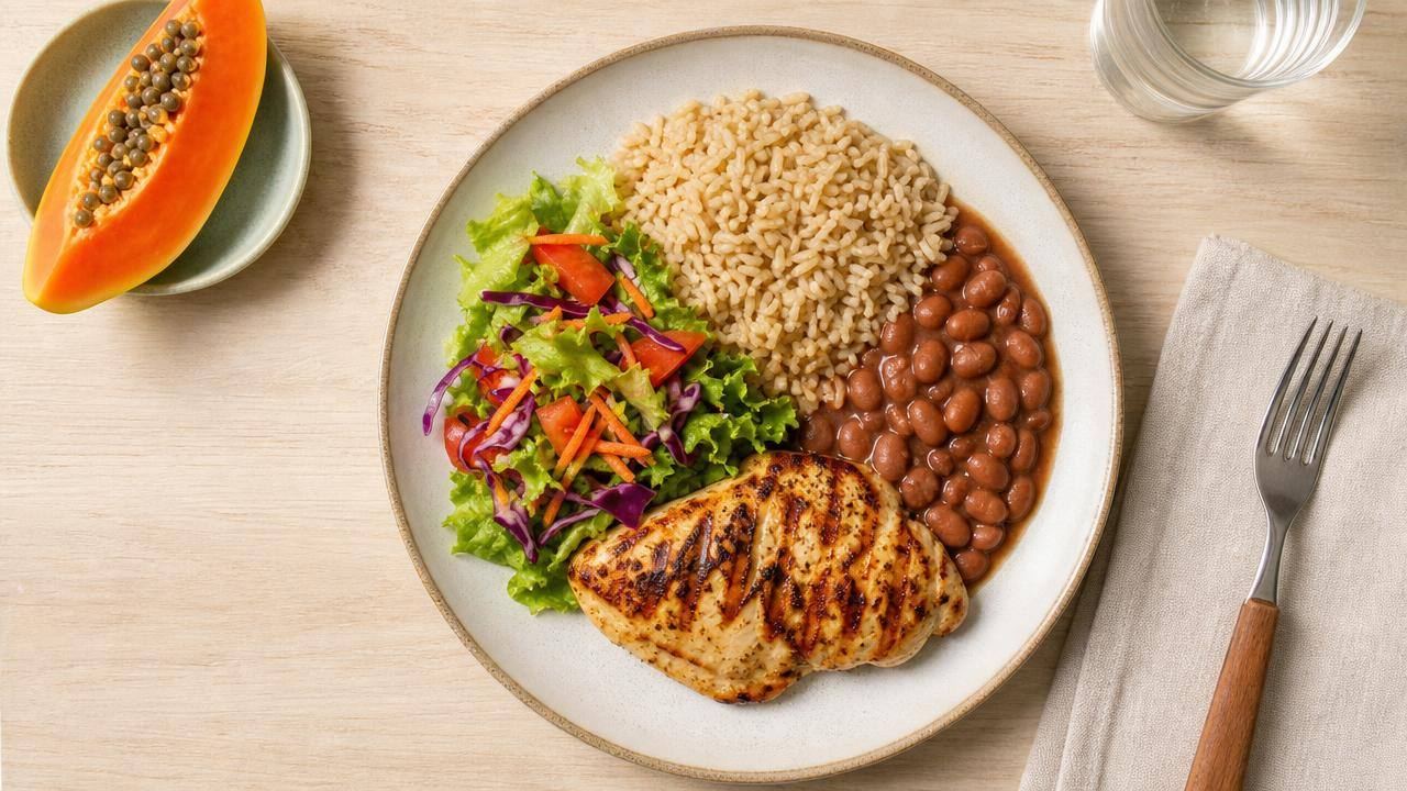 Prato equilibrado com arroz, feijão, frango grelhado, salada e frutas em uma mesa clara