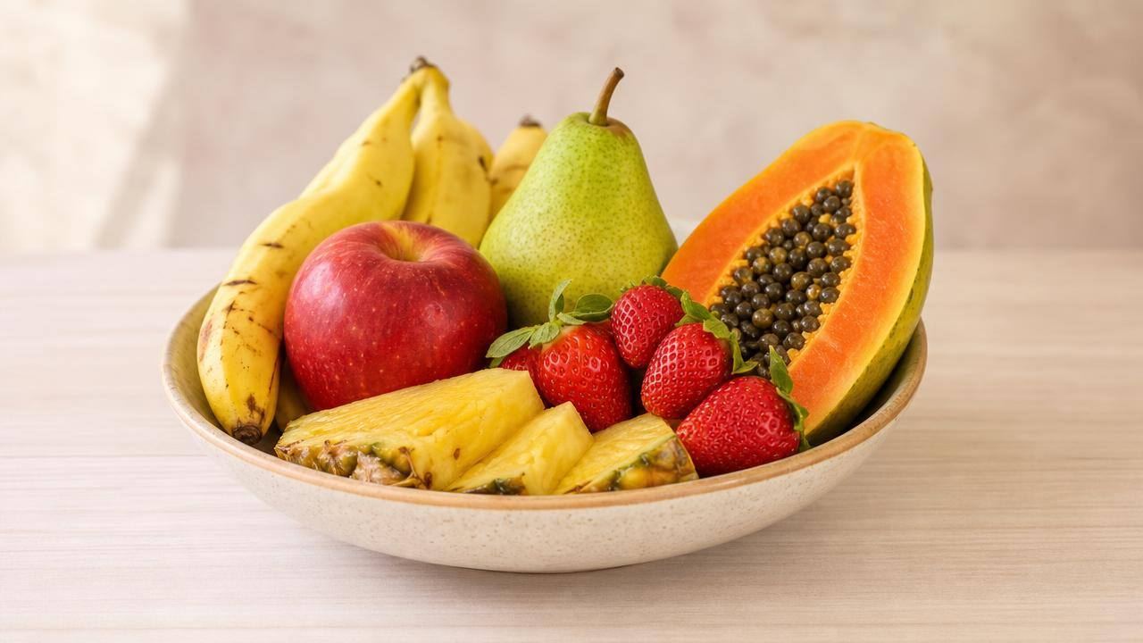Bowl centralizado com maçã, pera, mamão, morangos e banana sobre mesa clara, em composição editorial limpa.