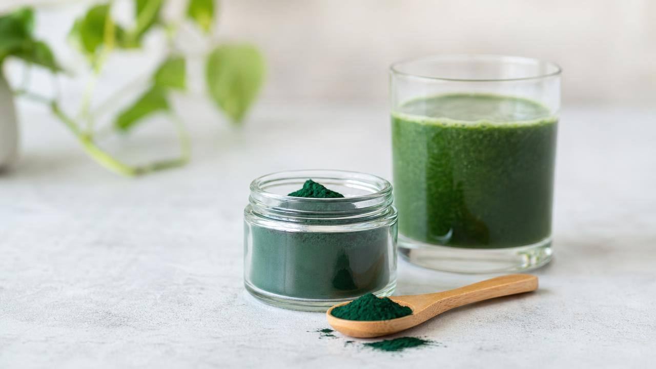 Pó verde de spirulina em uma colher de madeira ao lado de um copo com bebida verde sobre fundo claro