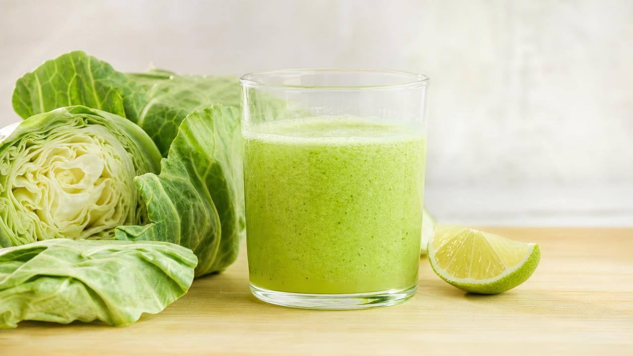 Copo de suco verde de repolho ao lado de folhas frescas de repolho sobre bancada clara