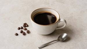 Xícara de café preto ao lado de grãos de café e uma pequena colher, em mesa clara e organizada