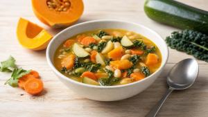 Tigela de sopa caseira com legumes variados sobre mesa clara, em composição editorial limpa