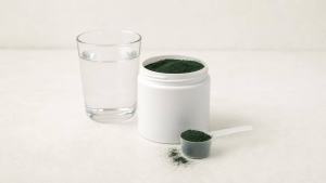 Pote de suplemento de spirulina em pó ao lado de colher medidora e copo com água, em fundo claro e limpo