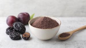 Tigela com farinha de ameixa ao lado de ameixas secas e colher de madeira em mesa clara